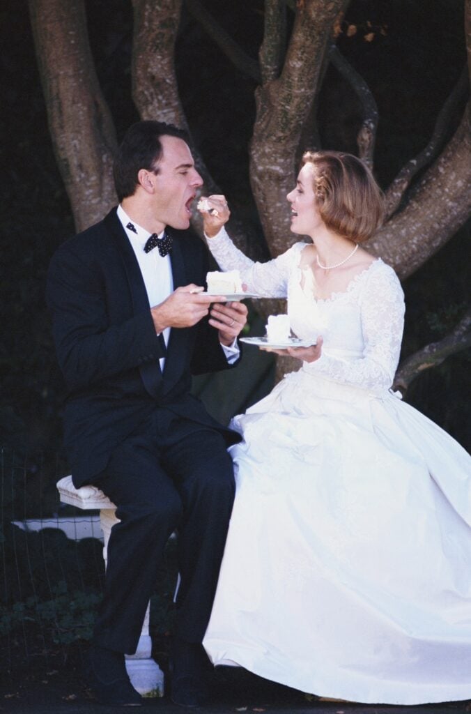 Bride feeding wedding cake to groom while seated outdoors beneath a large tree Gallery Image