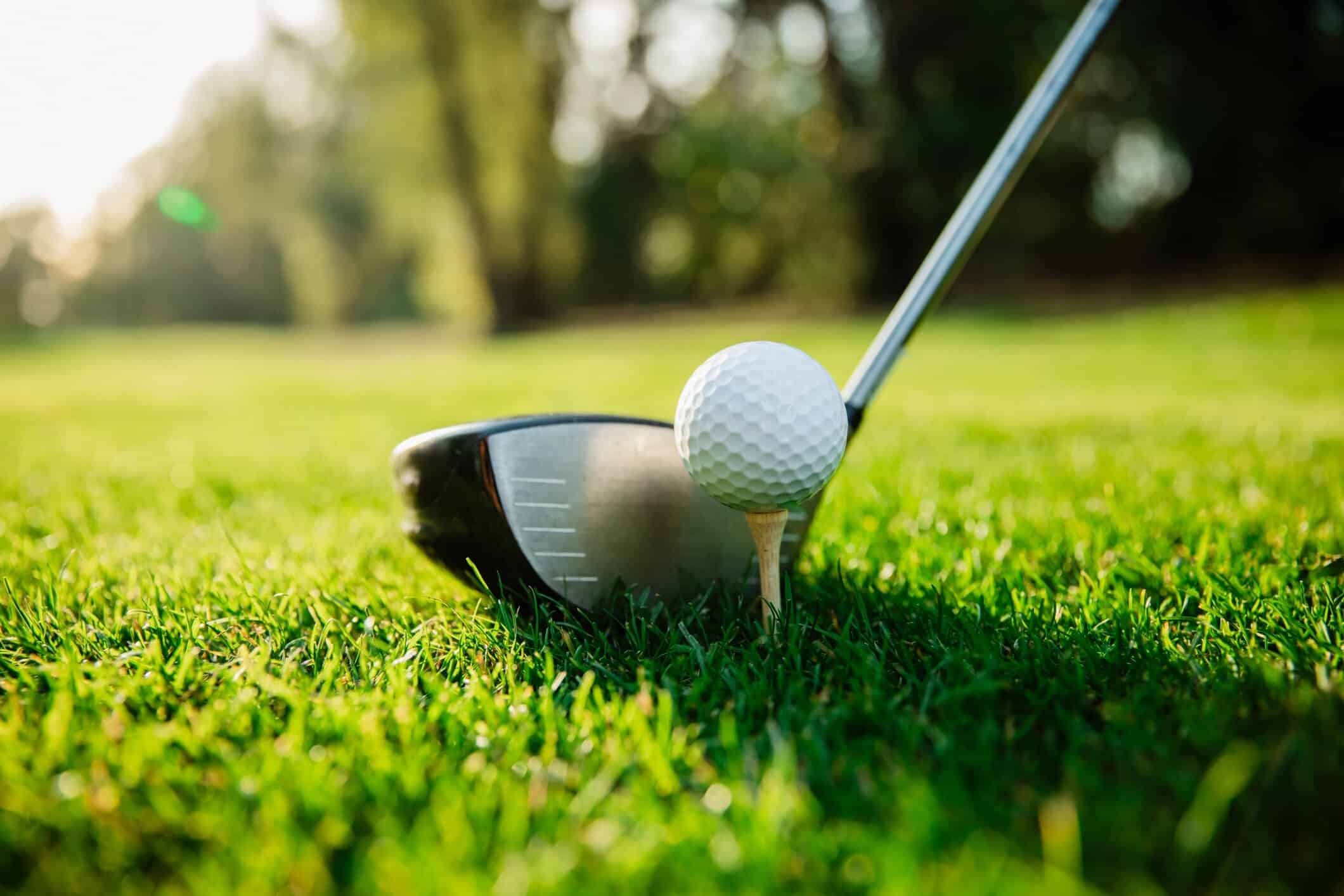 Close-up ground-level view of a golf ball on a tee with a driver club head on a green fairway
