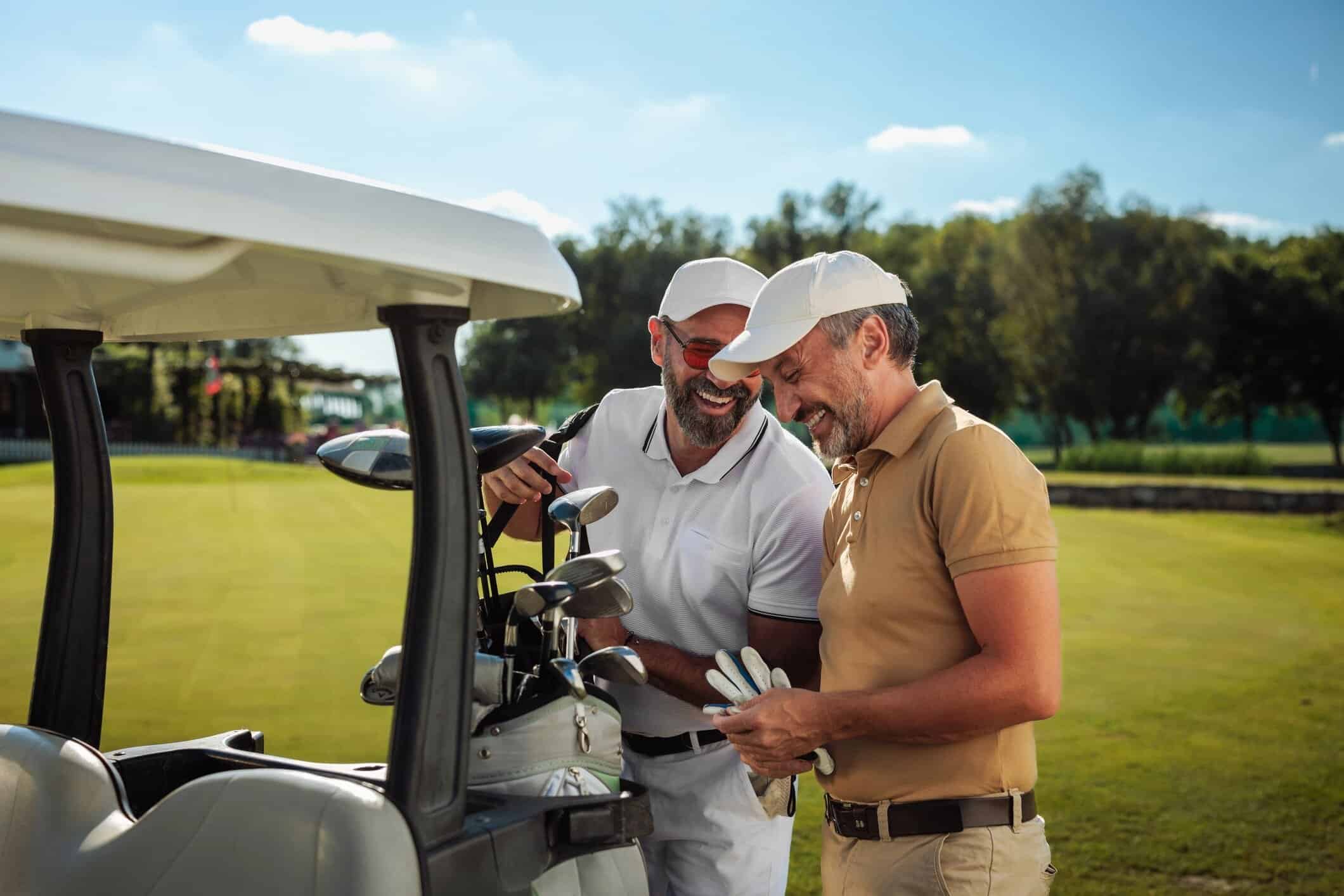 Two smiling men in golf caps laughing together beside a golf cart on a sunny course