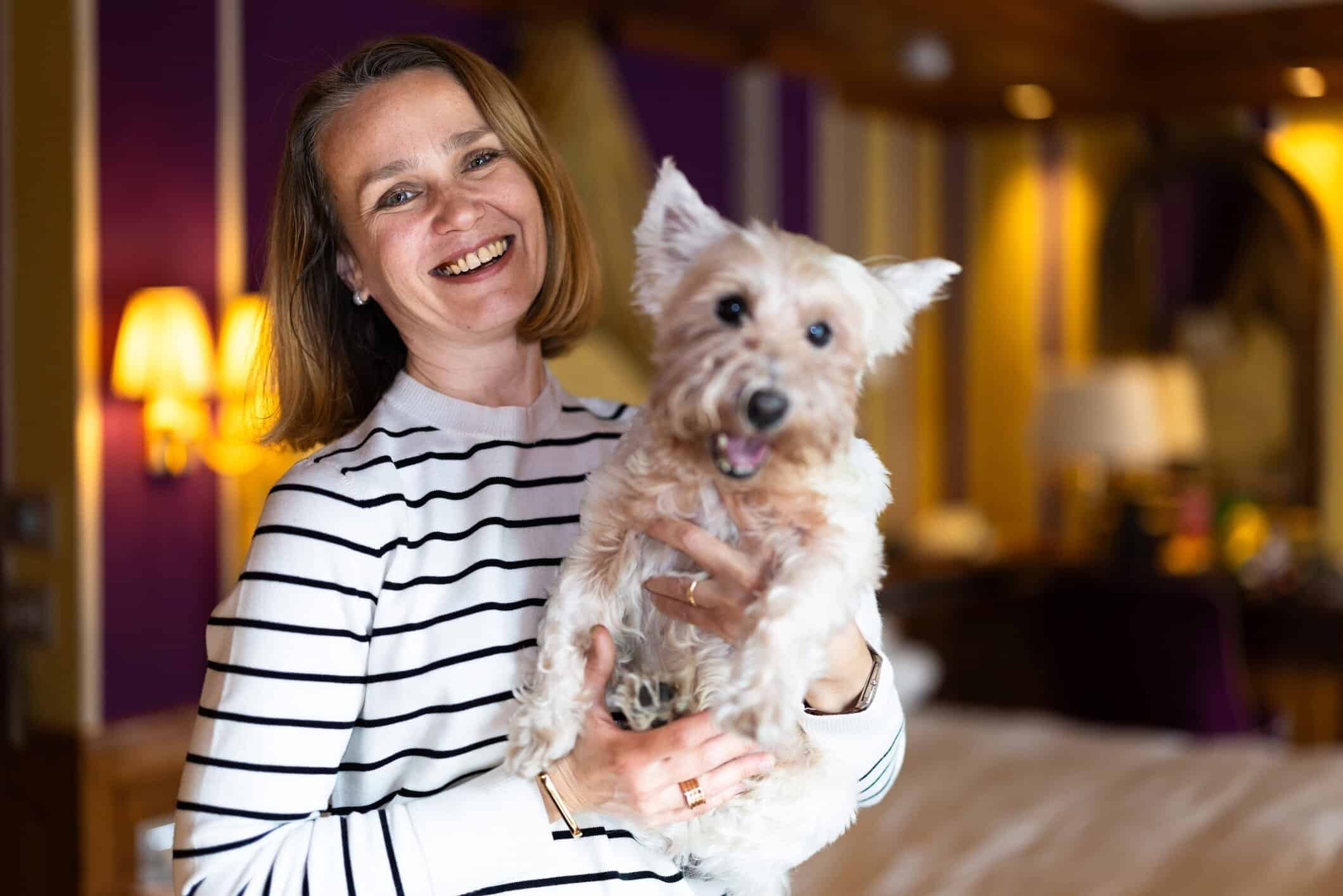 Smiling woman holding a fluffy white terrier dog inside a warmly lit hotel room