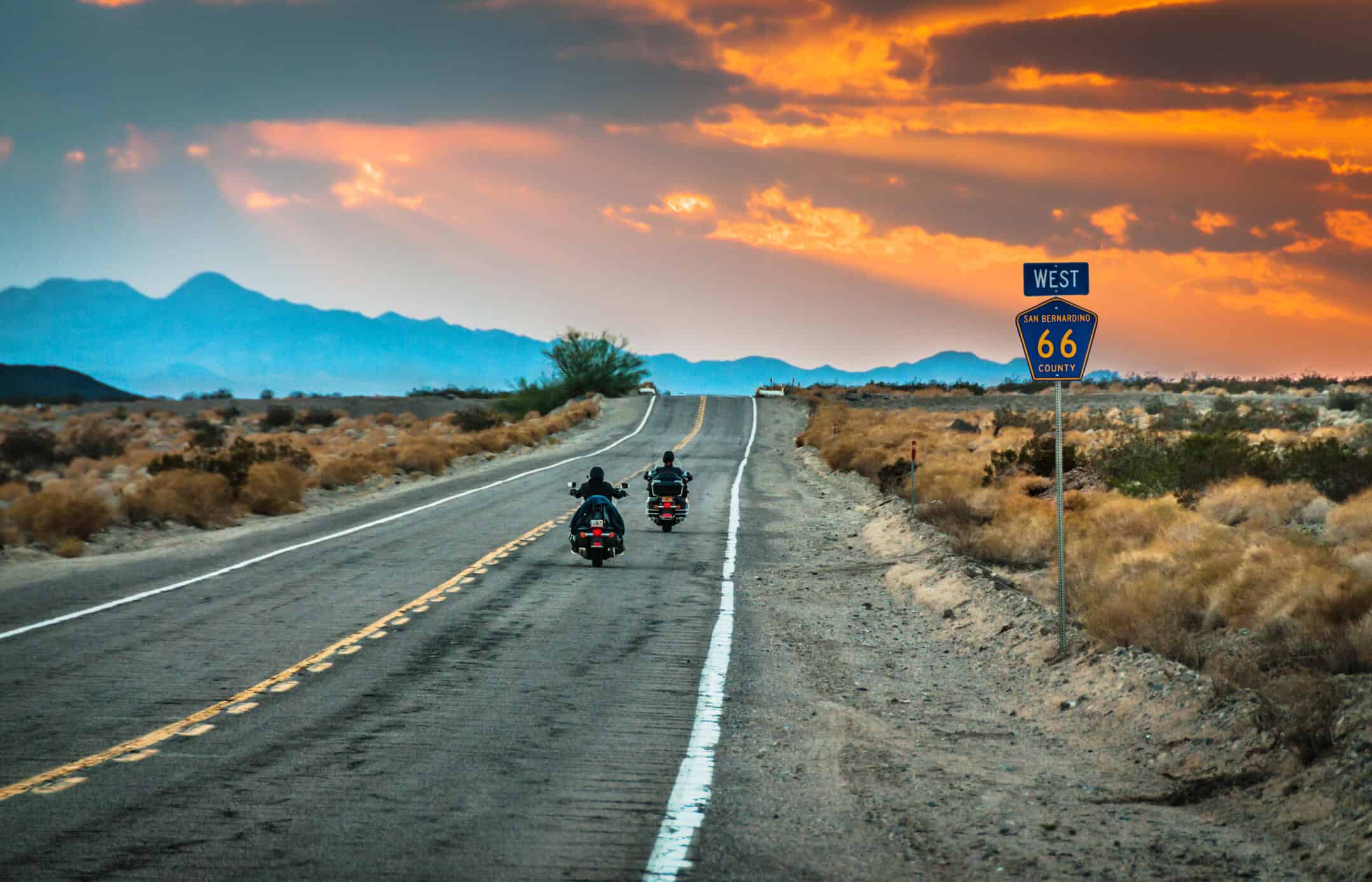 Two motorcyclists riding down a long desert highway at sunset along Route 66 in San Bernardino County