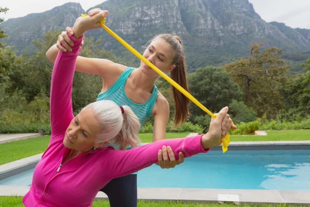 Front view of Caucasian female trainer assisting active senior Caucasian woman to exercise with resistance band in the backyard