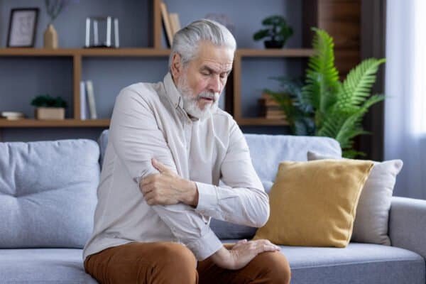 An elderly man experiencing discomfort holds his shoulder while seated on a couch, illustrating health issues in senior living.