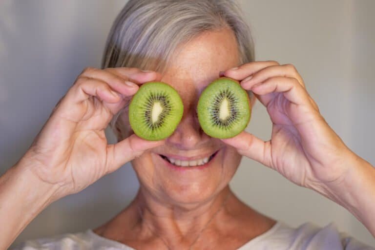 Elderly woman holding a ripe green kiwi cut in half in front of her eyes. Concept of fruit rich in vitamins and healthy eating.