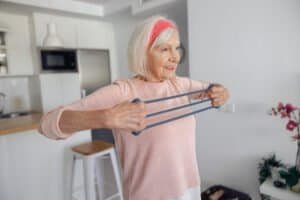 Happy senior lady strengthen her hands when doing workout with rubber expander on blurred background of apartment