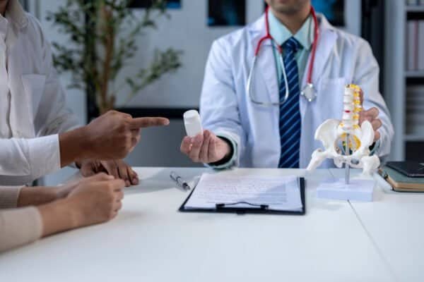 A doctor is holding a bottle of medicine and pointing to it. A woman is sitting at a table with the doctor and another person