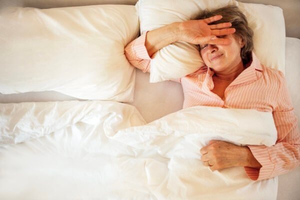 An elderly woman in pajamas is lying on a pillow in bed. Mature lady stretches and wakes up at home in the morning. The pensioner covers her hand from the light and smiles