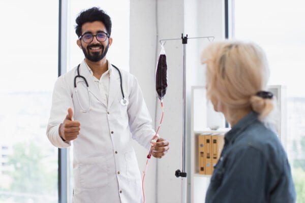 Experienced man in lab coat testing dropper with blood for transfusions near senior woman sitting on couch in exam room. Healthcare practitioner consulting female patient with cancer in clinic.