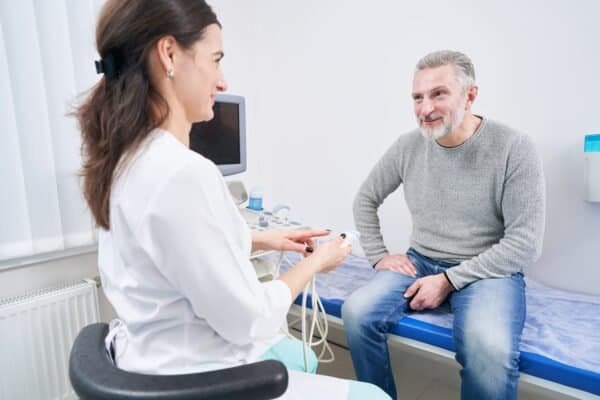 Smiling diagnostician demonstrating ultrasonic transducer to mature man seated on exam table
