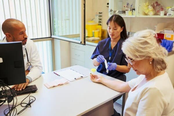 Nurse taking blood sample from finger of senior patient while doctor uses computer in modern hospital setting