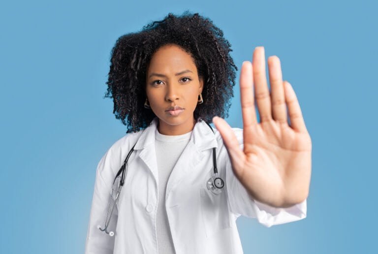 Serious confident young african american curly woman doctor therapist in white coat do stop sign with hand, isolated on blue background, studio. End of problems, fight against disease, health care