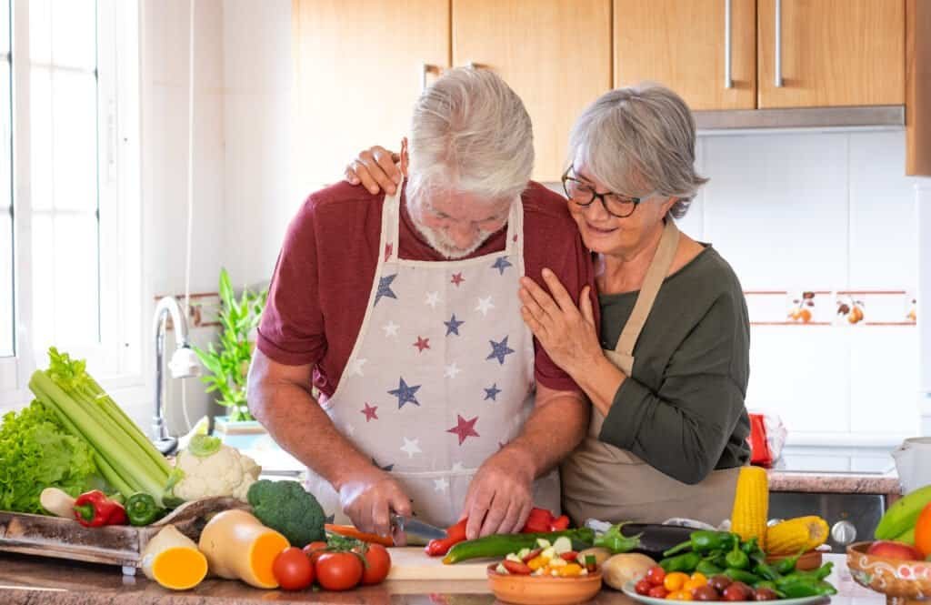 Vegetarian lifestyle. Beautiful elderly couple white-haired hug in the kitchen preparing a vegetable soup. On the table a mix the of raw seasonal vegetables
