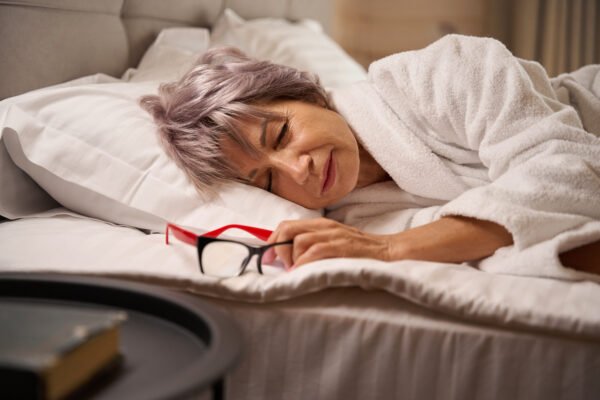 Woman is dozing in a bright bedroom on soft pillows, reading glasses nearby