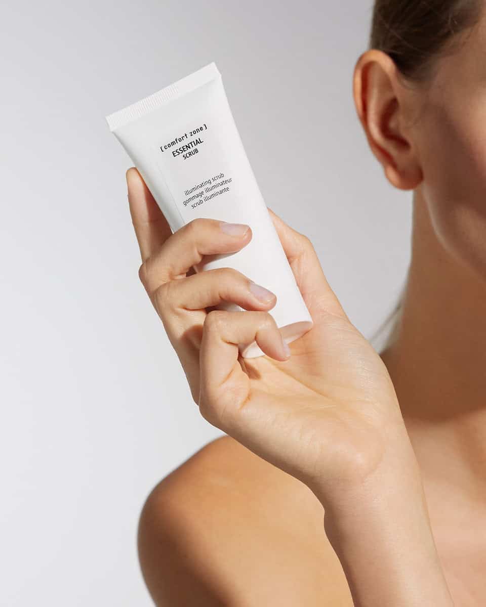 Woman holding a white tube of essential body scrub for skin exfoliation.