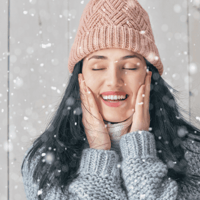 Beautiful woman in winter hat and sweater enjoying snow, promoting winter skincare routines.