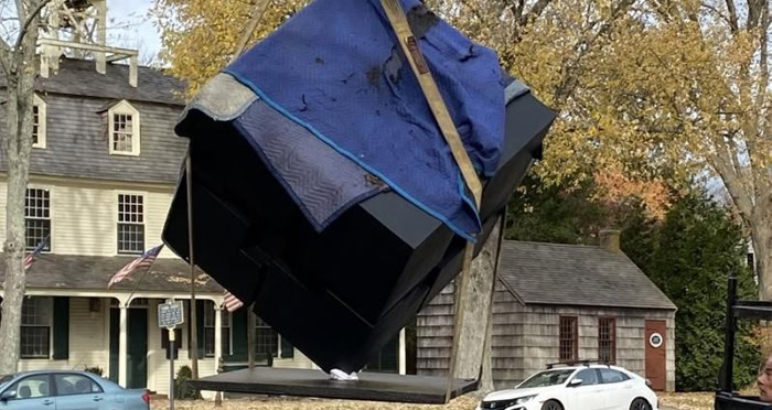 Suspended blue blanket covering a large black object outdoors in a park with autumn trees and historic buildings in the background.