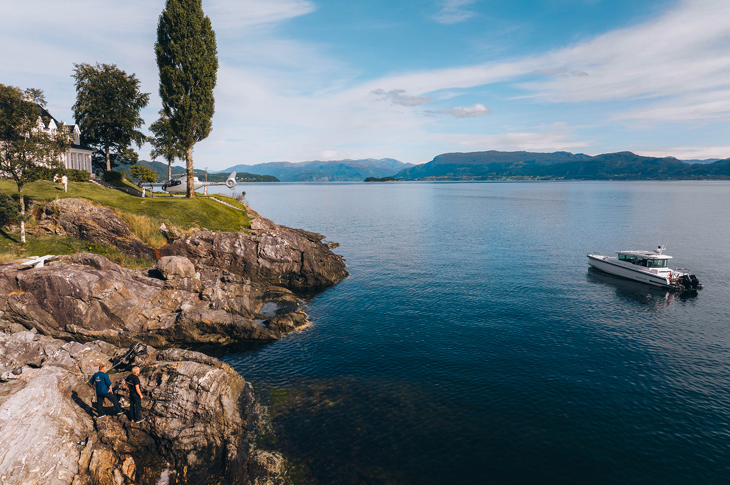 Seaside waterfront with luxury home, rocky shoreline, and yacht cruising in scenic fjord landscape.