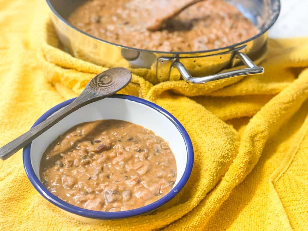 Vegan refried beans in small white enamel bowl with blue rim, wooden spoon, and cooking skillet to side, all sitting on yellow tea towel.