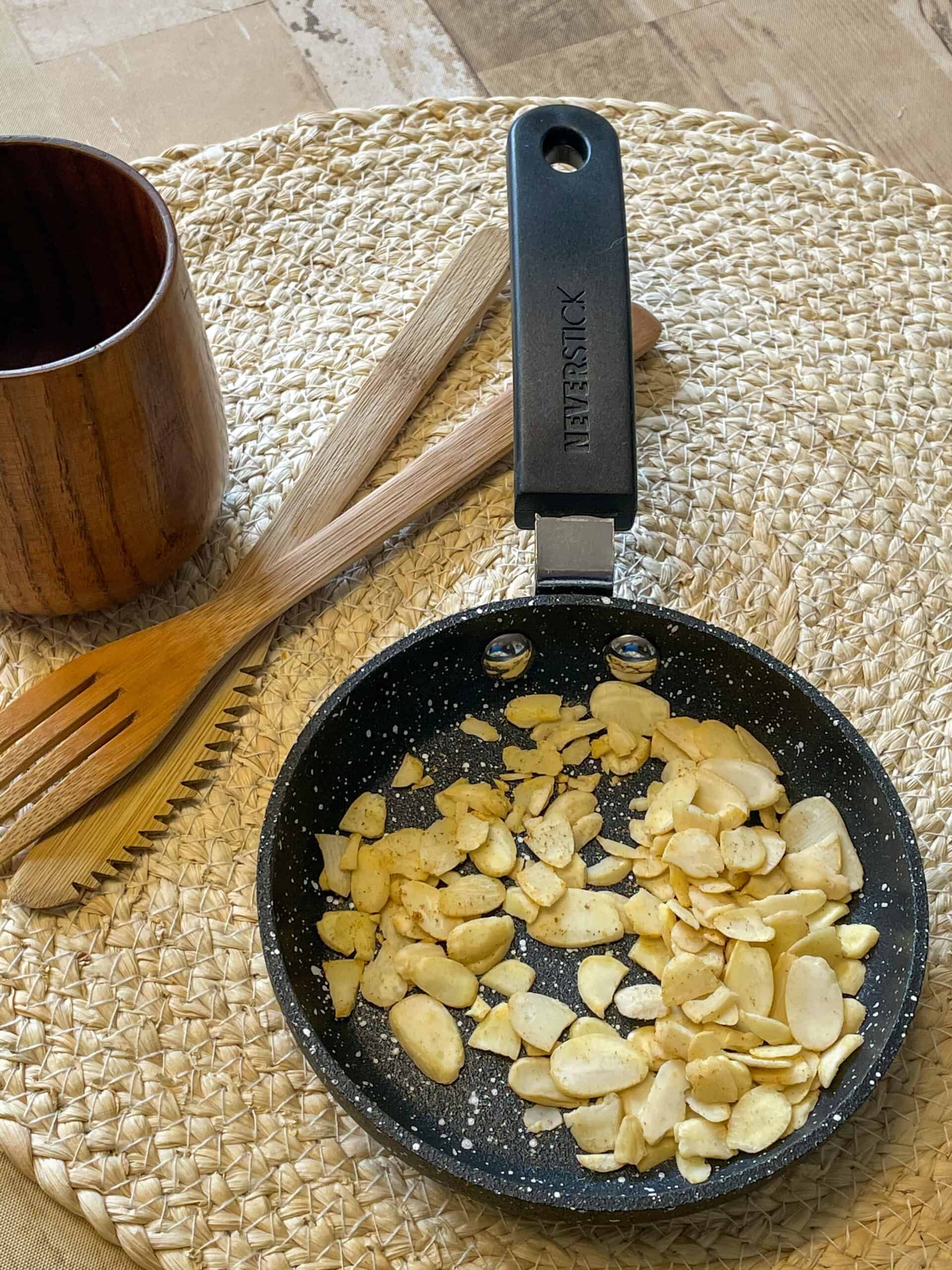 Toasted almonds in a small pan on weaved mat, wooden fork and spoon, small wooden cup.