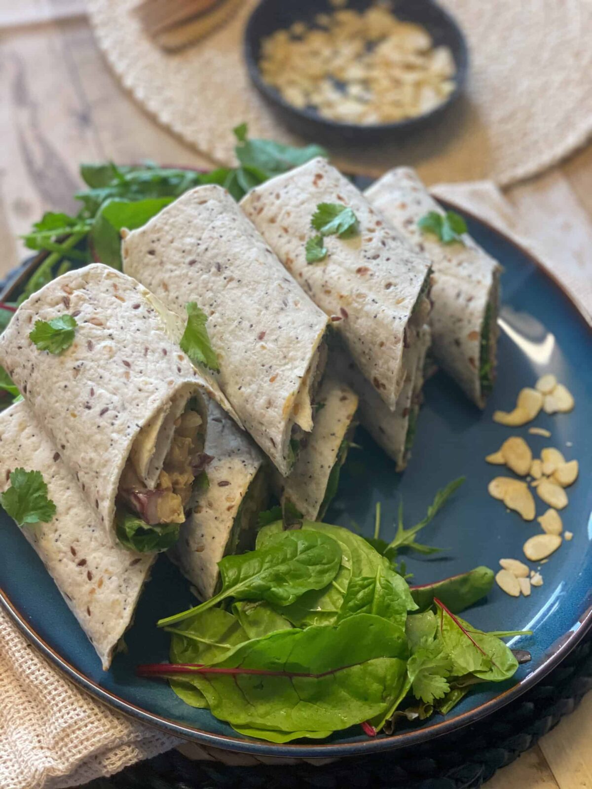 A stack of tortilla wraps filled with Coronation chickpea filling, sitting on blue plate with extra side salad and sprinkle of flaked almonds.