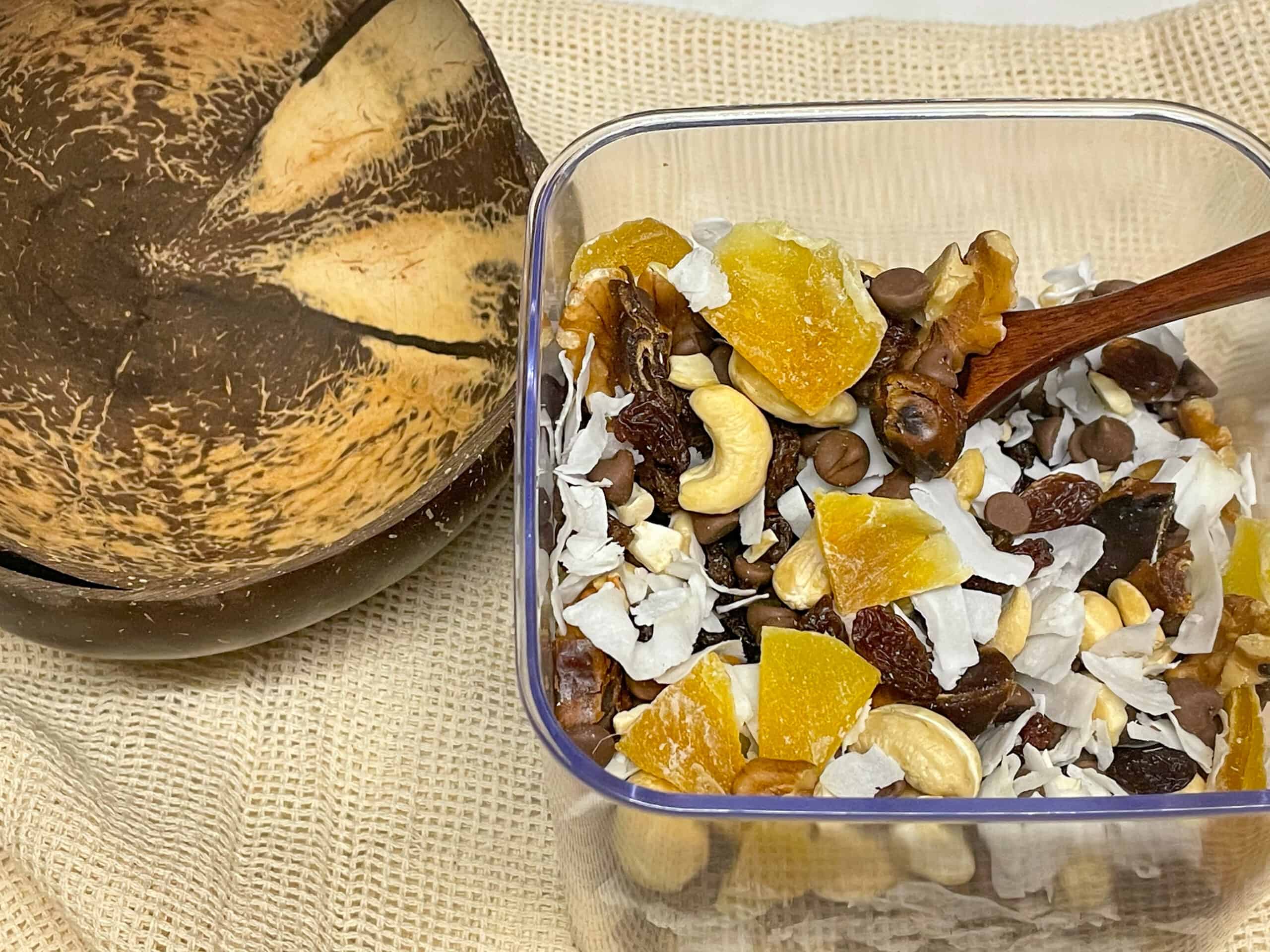 Trail mix in a food container with wooden spoon in container, a coconut bowl to side.