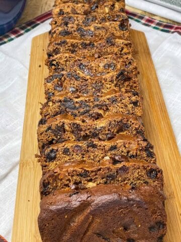 Fruit loaf cake sliced into portions on a wooden cutting board with white linen table mat with tartan trim.