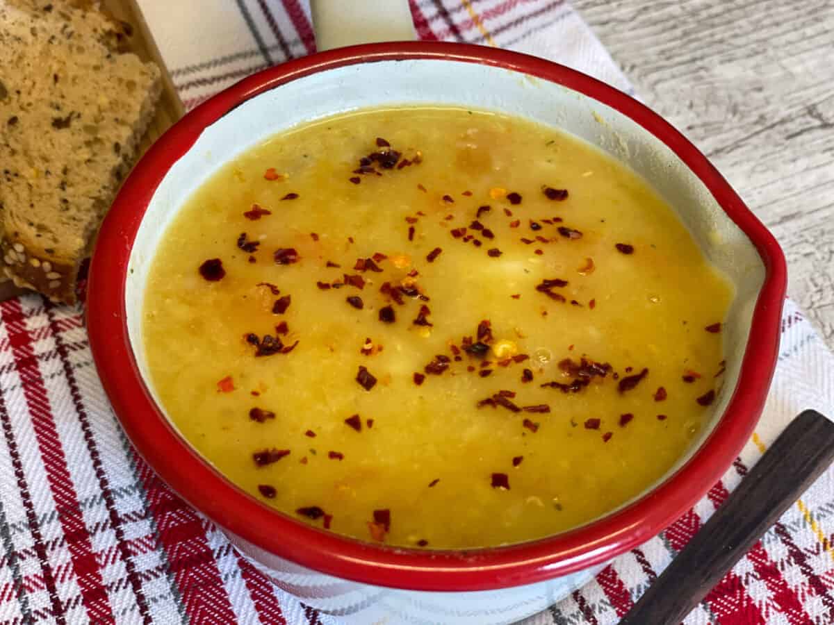 Lentil soup with red pepper flakes garnish, pieces of brown bread, wooden spoon to side and red check tea towel underneath.