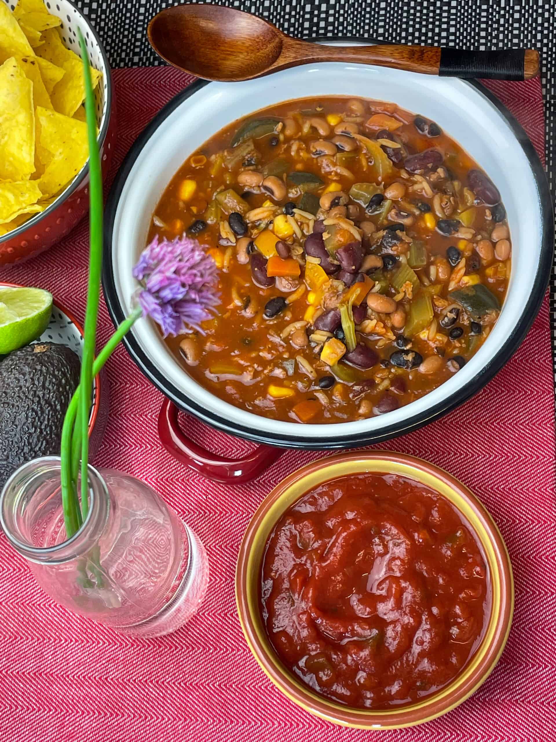 Taco bean soup with small bowl of salsa, tortilla chips off to the side, and a chive flower in a glass bottle. Red background.