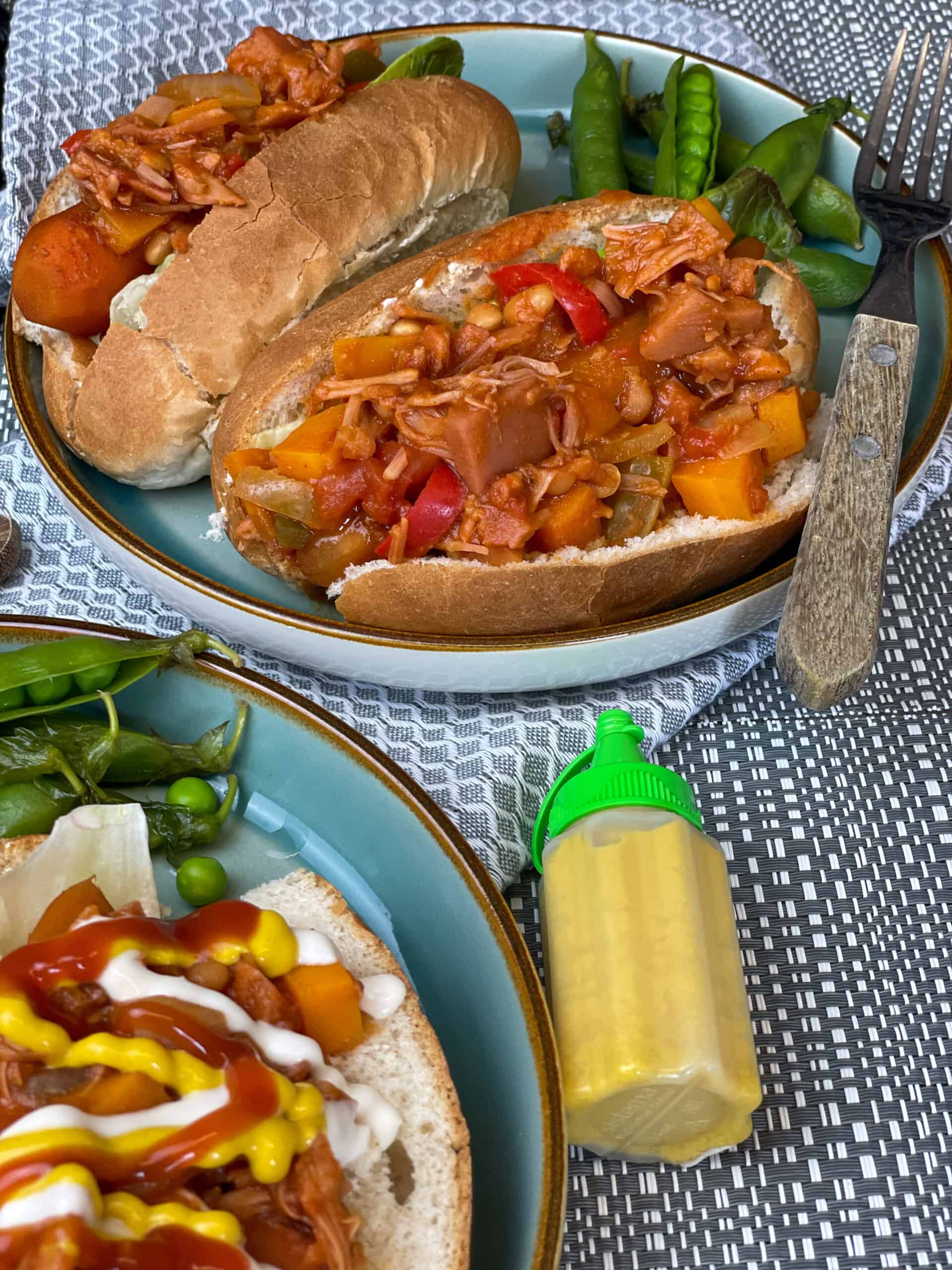 Carrot hotdogs in rolls with campfire stew topping, mustard bottle to side, blue background.