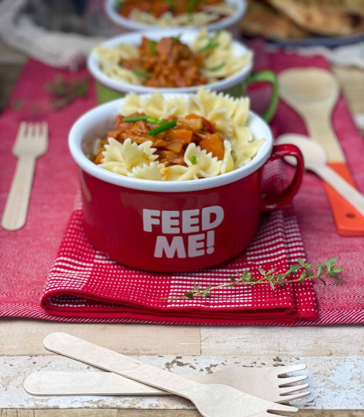 Large red mug with 'feed me' text, forks to side, garlic bread in distance, red background.