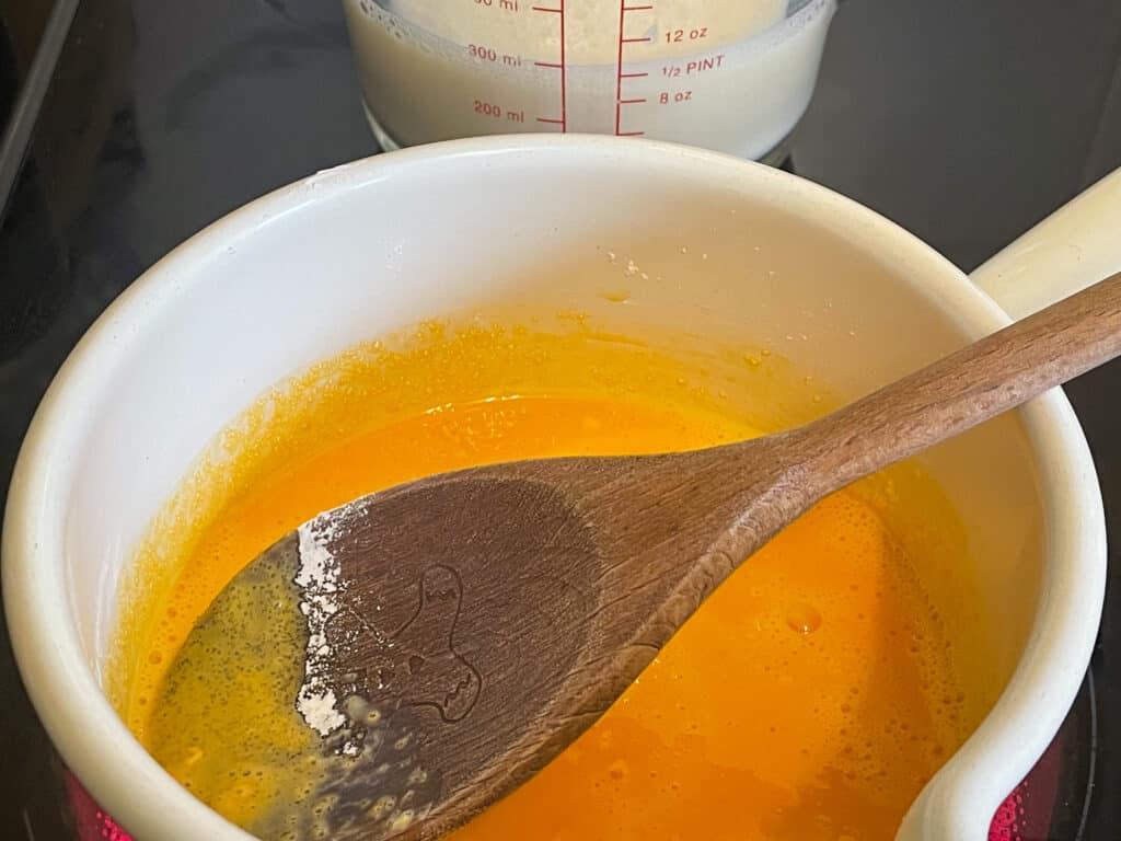 Custard being cooked in small cream pan with wooden spoon, milk in jug off to the side.