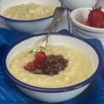 vegan slow cooker rice pudding served in enamel white bowls with small bowl of strawberries, and pot of jam in distance, blue background.