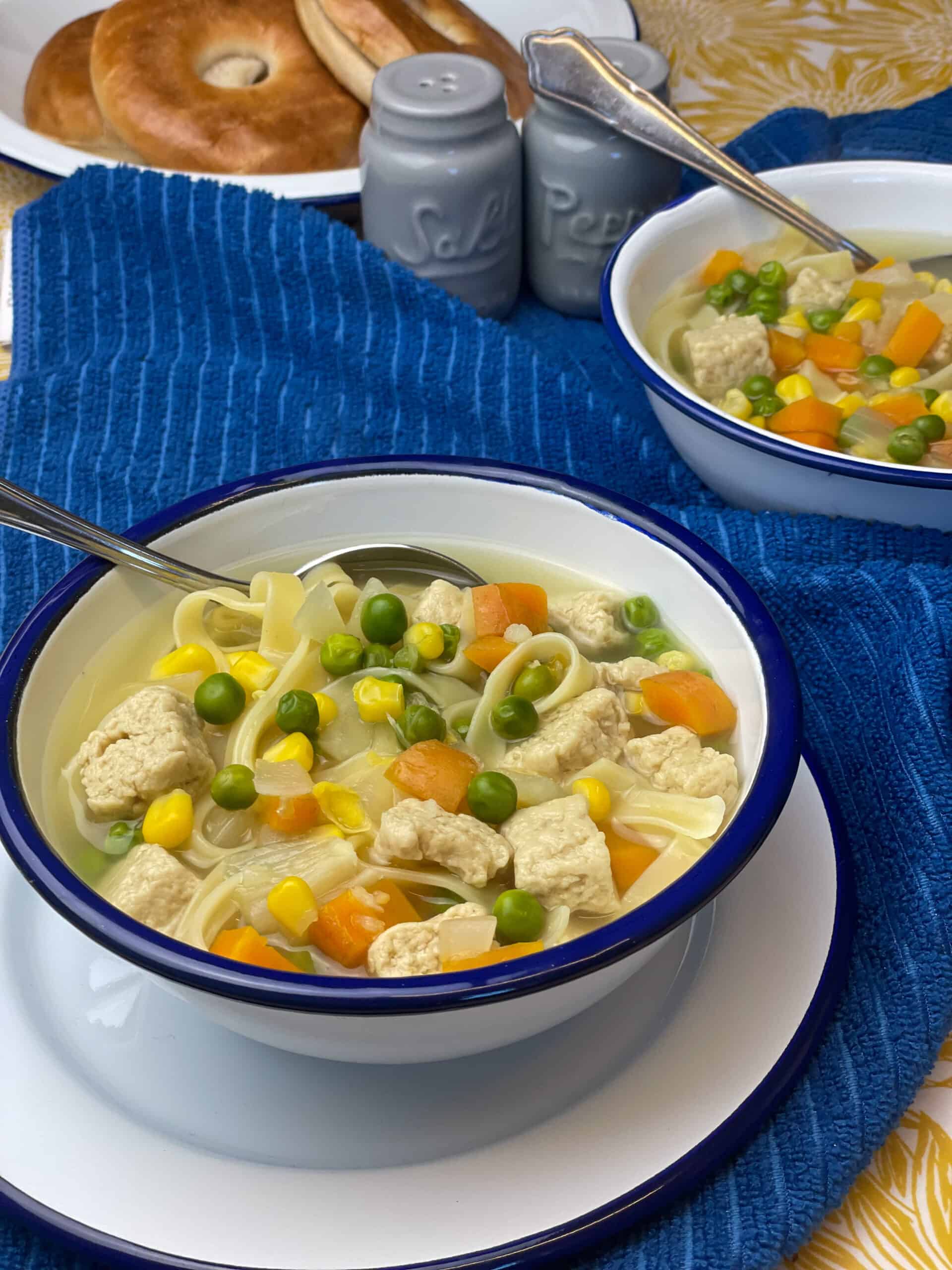 A bowl of soup with a second bowl in the background, with a blue tea towel background and a plate of bagels in background.