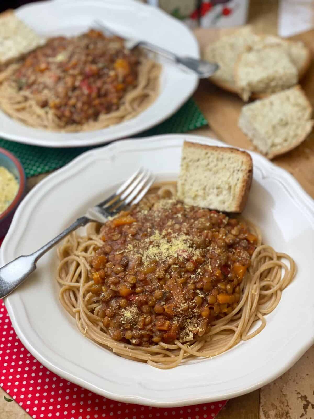Two bowls of slow cooker lentil spaghetti Bolognese, with sprinkle of nutritional yeast, wedge of her bread, silver fork and red check tea towel, featured image.