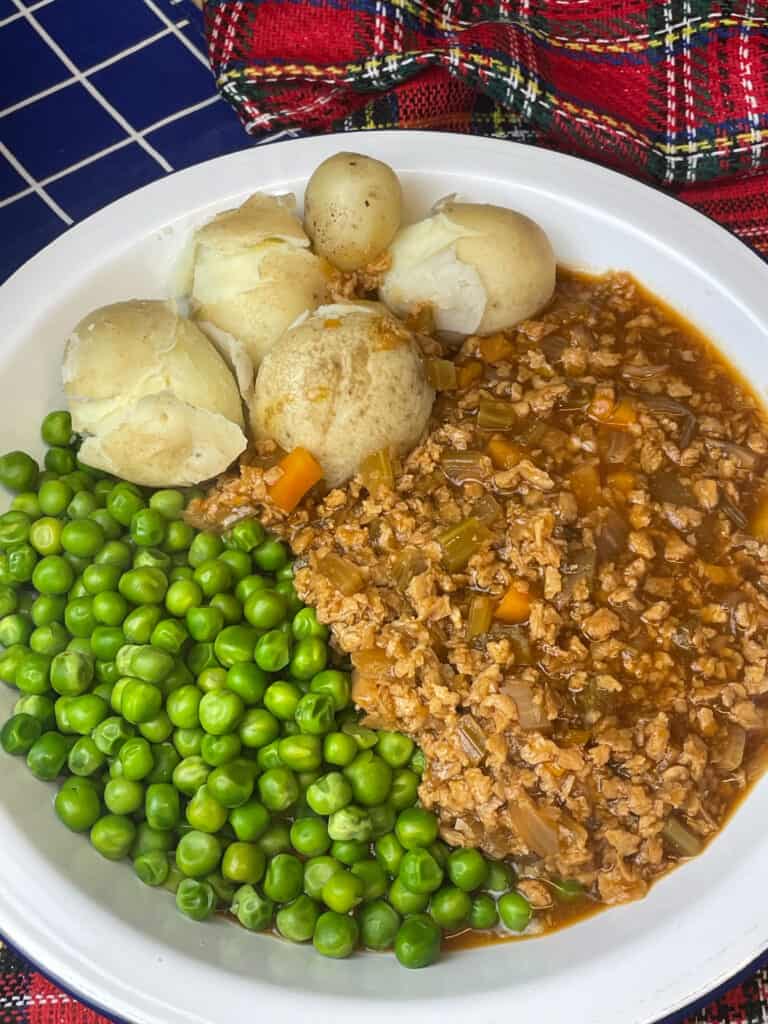 plate of vegan mince and tatties, with peas, tartan background and small blue white check tea towel to side.