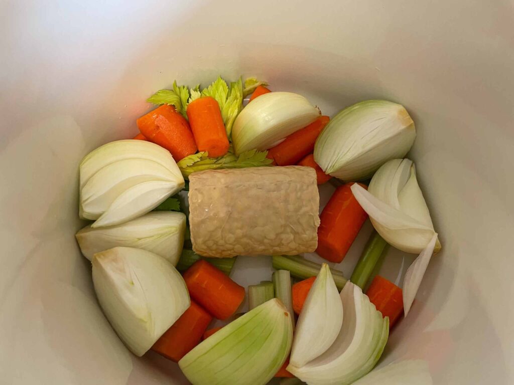 Tempeh nestled in a stock pot with onion wedges, carrot slices, celery stalks.