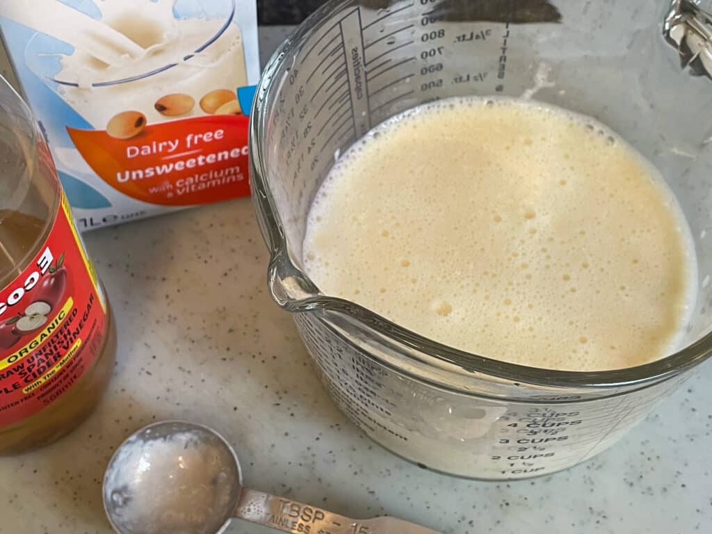 Glass jug of soya milk, with bottle of apple cider vinegar and carton of soya milk to side, with measuring spoon, white chopping board background.