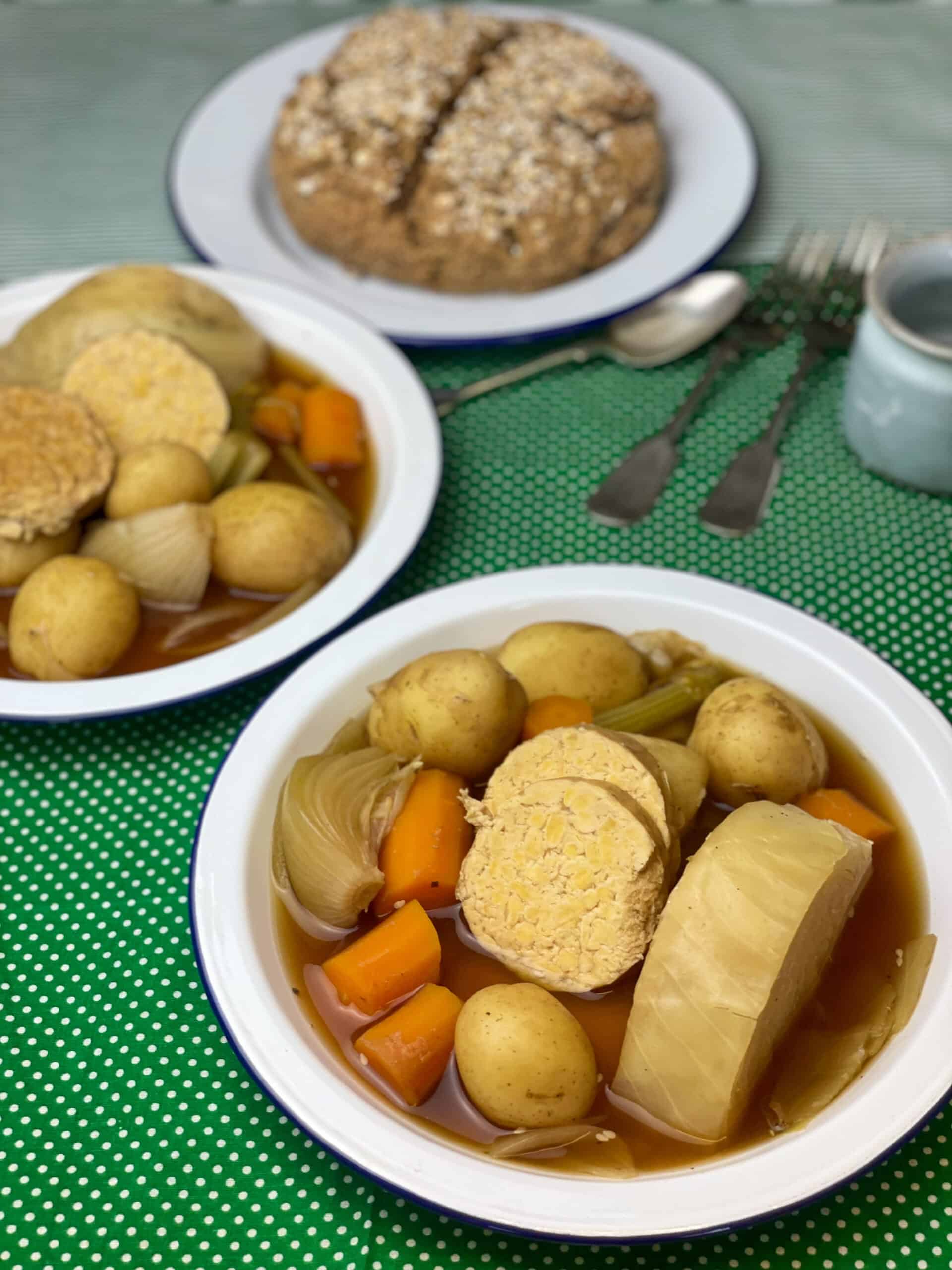 Two plates of vegan corned beef and cabbage stew with plate of soda bread, forks and spoons to side, small blue mug to side and green polka dot background.