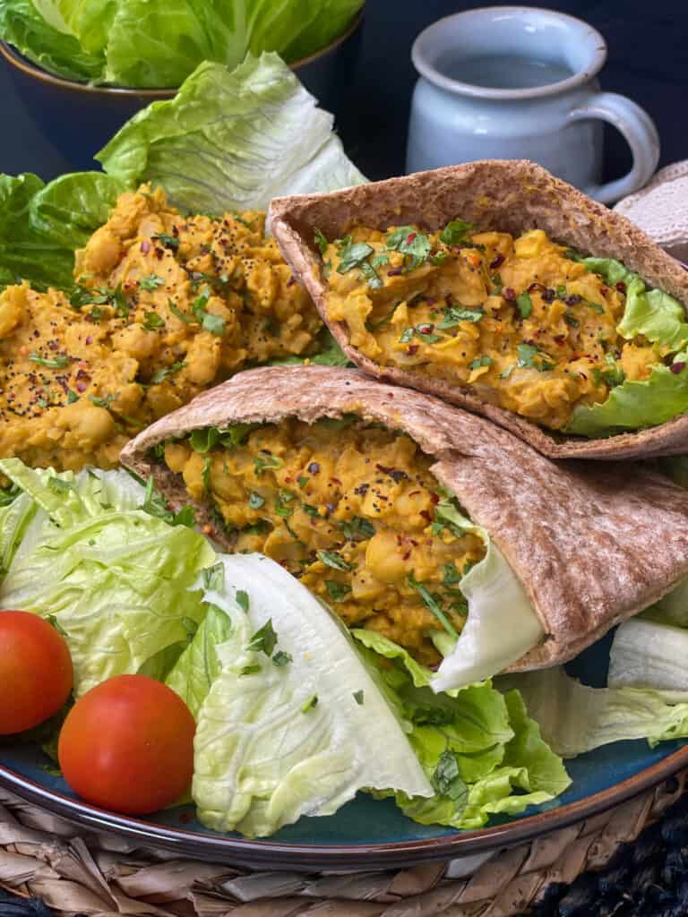 stuffed pitta breads with scrambled chickpeas and lettuce, single romaine leaf with scramble, cherry tomatoes at side of large blue plate, straw mat underneath and small blue cup to side.