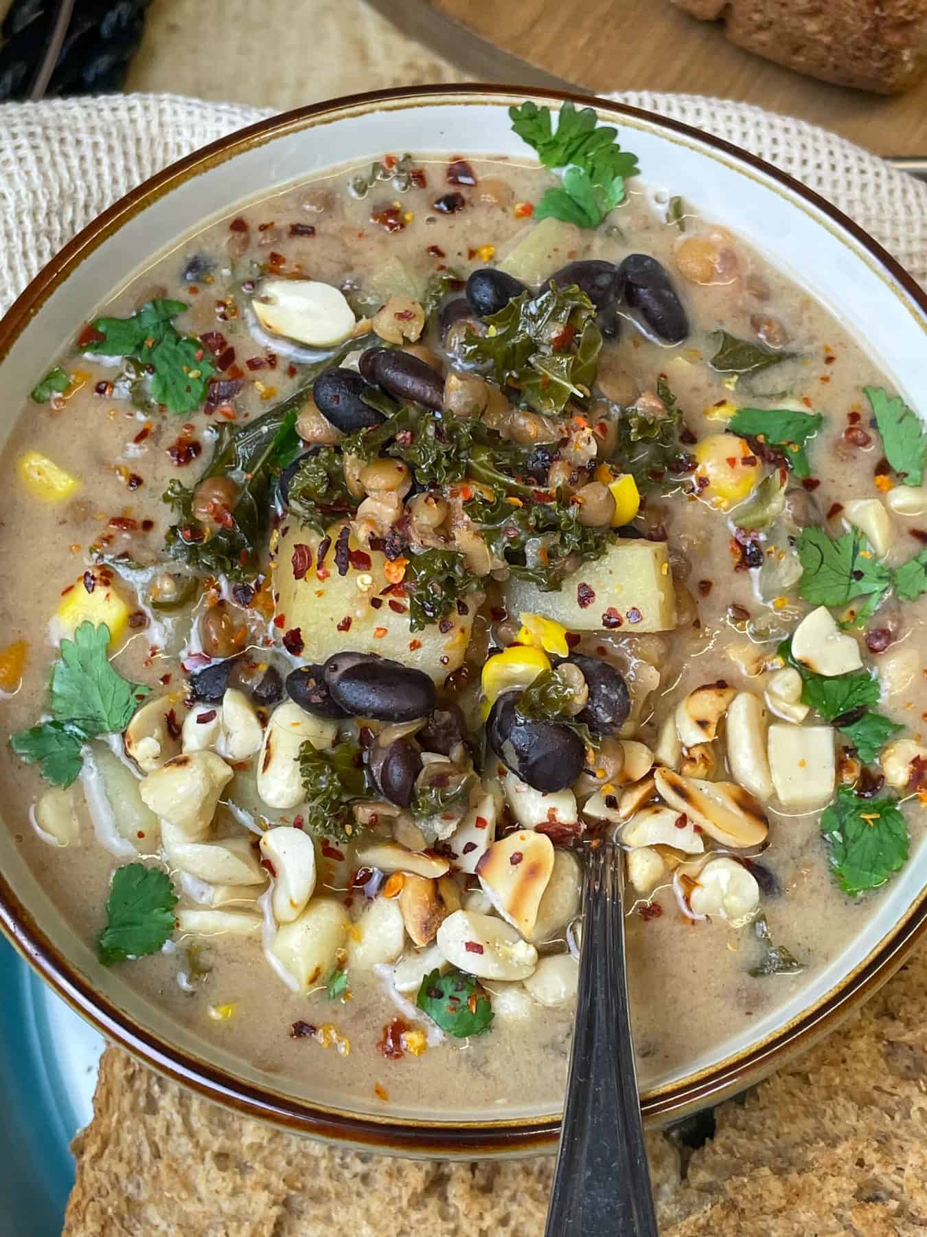 A close up of slow cooker spicy peanut soup in bowl with brown rim, bread to side, silver spoon.