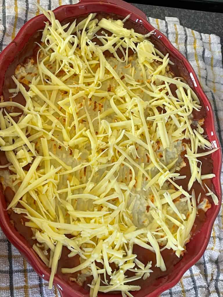 Grated vegan cheese sprinkled over baked cowboy pie, sitting on yellow and grey tea towel.
