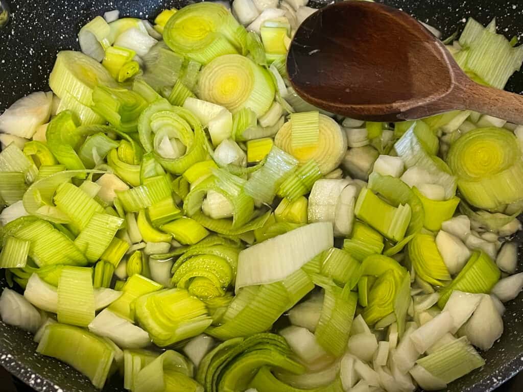 Leeks, onions and garlic cooking in pan with wooden spoon to side.
