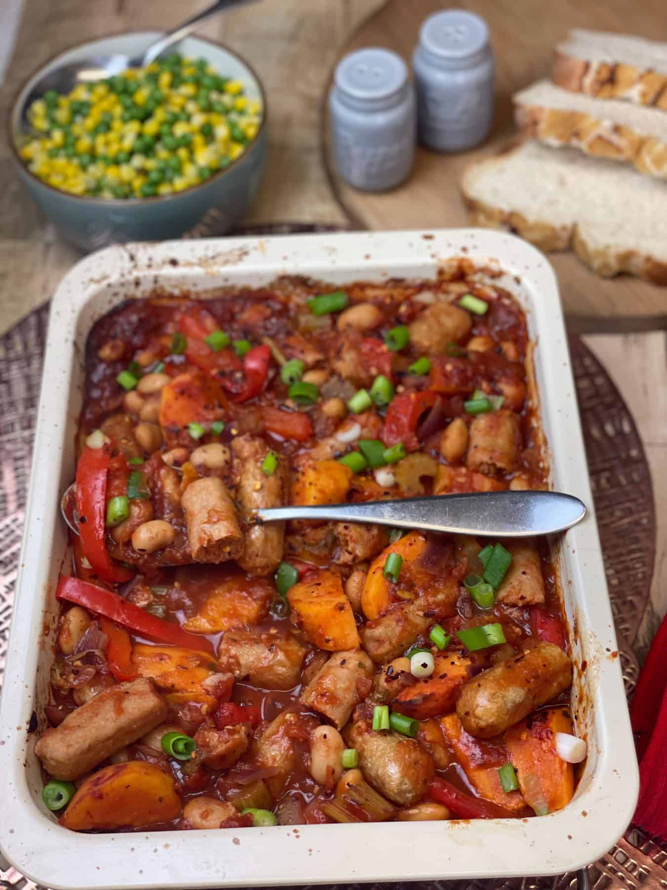 Rectangular casserole tray with veggie sausage casserole and silver serving spoon, bowl of peas and corn to top, along with blue salt and pepper shakers and three slices of white crusty bread on wooden board.