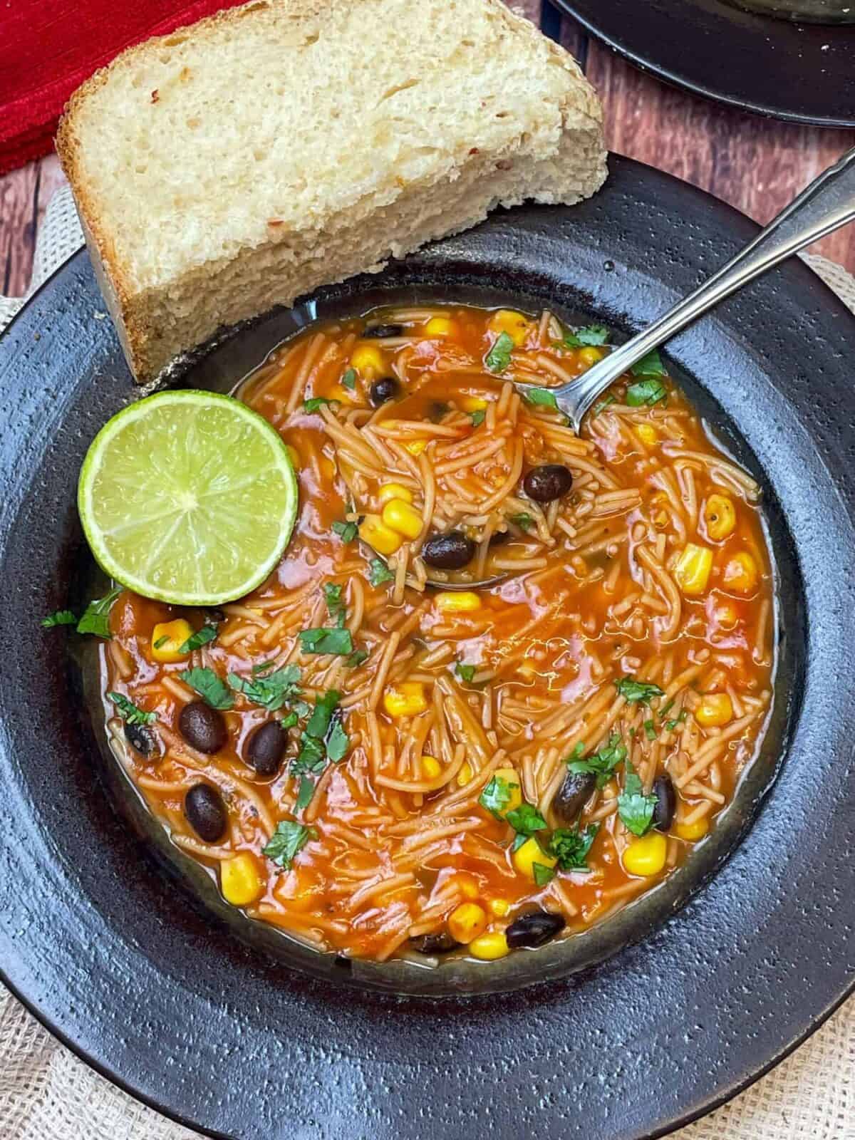 A dark brown clay bowl with Mexican noodle soup, lime wedge and silver spoon in bowl, and wedge of crusty bread to side.