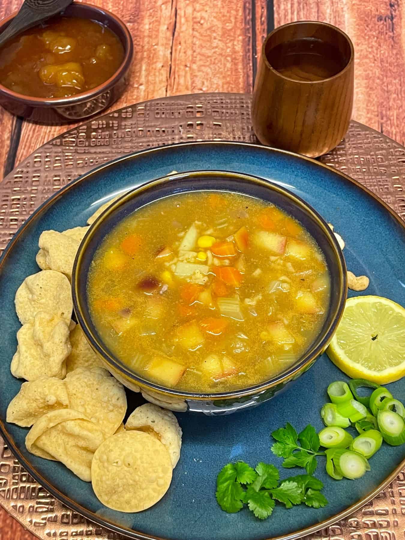 Curried soup served in blue bowl on blue plate, with wooden cup to side and wooden bowl with mango chutney, wooden background and metallic mat.