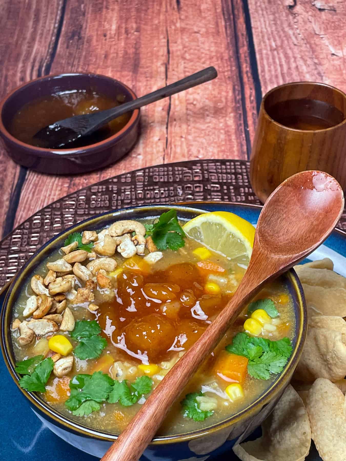 Blue bowl of Mulligatawny soup with wooden spoon, wooden cup, small dish of mango chutney and brown wood background.
