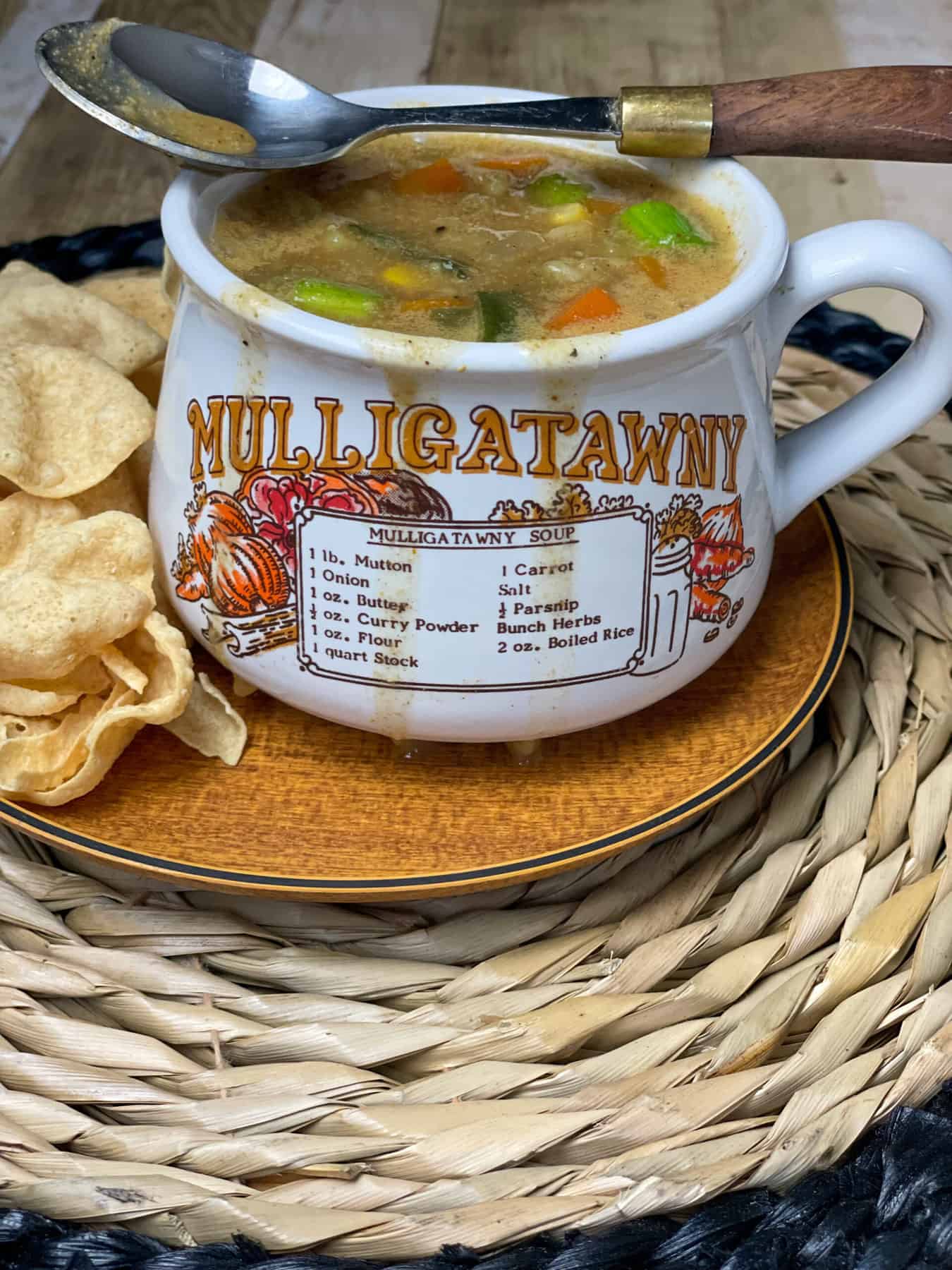 A soup mug full of soup, with the word Mulligatawny written on the mug with illustrations and recipe, served on wooden plate with mini poppadum's and wicker mat.