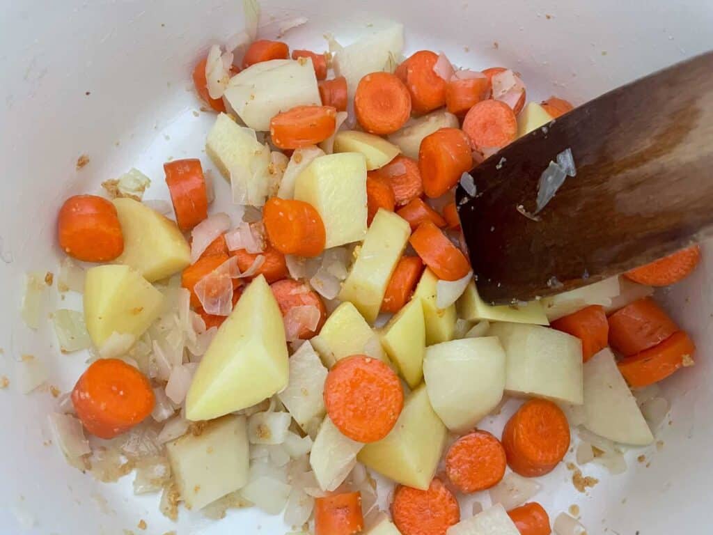 Carrot and potatoes added to soup pan to cook, wooden spatula to side.