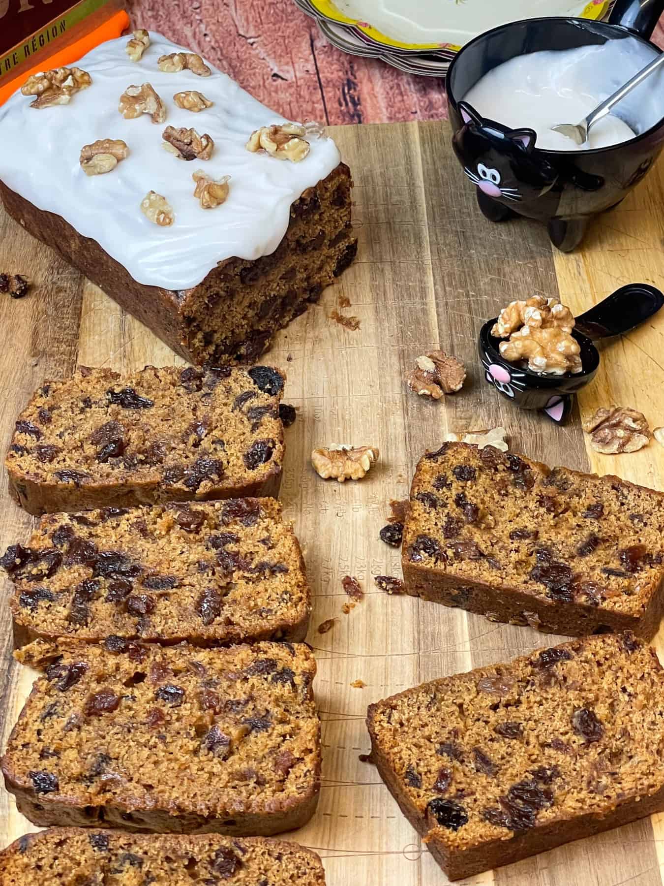 Slices of pumpkin fruit cake with large piece covered with cream cheese frosting and walnuts, wooden chopping board background, cat themed measuring set.