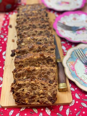 Whole fruit cake sliced into pieces on a cutting board, with paper party plates to side.